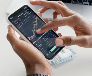 Hands using smartphone trading app with charts, dollar bills in background.